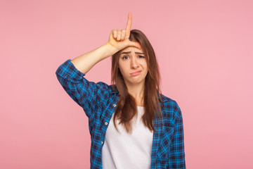 Portrait of depressed unlucky girl in checkered shirt showing loser gesture with fingers on...