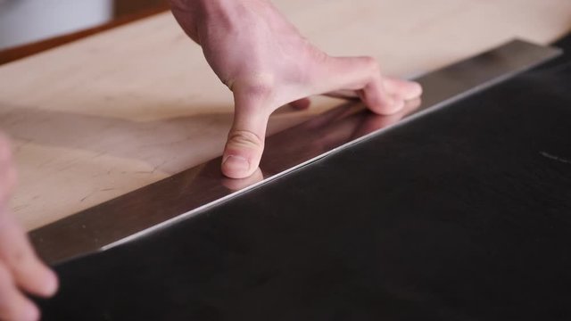 A tanner is cutting black leather sheet with a razor and metre. The master is making a leather belt for men.