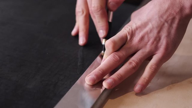 A tanner is cutting black leather sheet with a razor and metre. The master is making a leather belt for men.