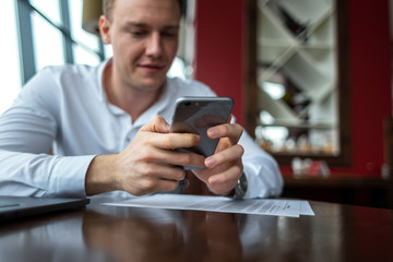 Young businessman wearing a white fashion shirt chatting and talking business on a mobile cell phone in a cafe with a laptop and documents. Freelance and selfemployment concept. Distance job.