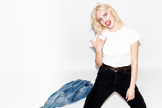 young stylish blonde girl in a white t-shirt and black jeans shows a rock sign on a white background, studio fashion photo with copy space