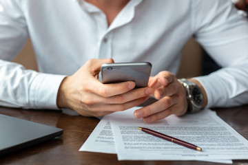 Closeup of hands of young businessman wearing a white fashion shirt with a mobile phone in a cafe with a laptop and documents. Freelance and selfemployment concept. Distance job.