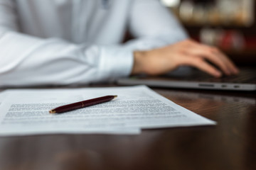 Fototapeta premium Closeup of documents and pen on the table. Young businessman wearing a white fashion shirt in a cafe with a laptop on the background. Freelance and selfemployment concept. Distance job.