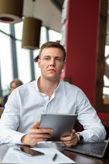 Young businessman wearing a white fashion shirt with a tablet in a cafe with a laptop, phone and documents. Freelance and selfemployment concept. Distance job.