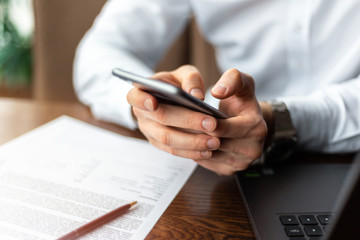 Fototapeta premium Closeup of hands of young businessman wearing a white fashion shirt with a mobile phone in a cafe with a laptop and documents. Freelance and selfemployment concept. Distance job.