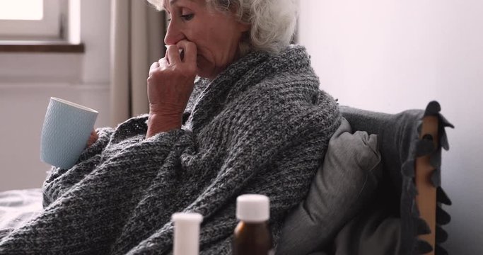 Older ill woman holding handkerchief blowing nose sitting in bed at home with cold remedy. Sick elder lady got flu virus having grippe symptom. Seniors respiratory diseases and antiviral drugs concept