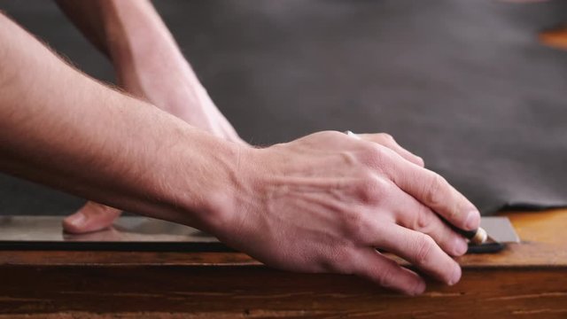 A tanner is cutting black leather sheet with a razor and metre. The master is making a leather belt for men.