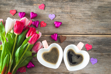 Holiday and celebration composition. Two mugs shape heart of coffee, chocolates and bunch of purple tulips on old non paint wooden background; Space for text, top view