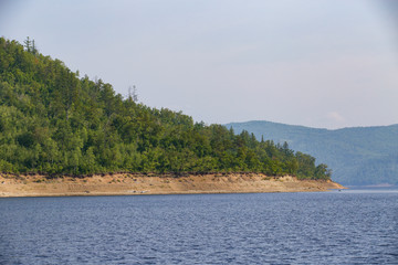 Beautiful landscape. Zeya reservoir, Amur region. A beautiful rocky shore with tall green trees goes into the calm blue waters of the reservoir.