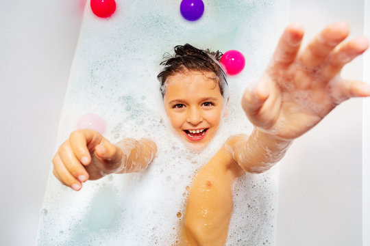 Cute boy with smile and laugh swim on the back in soap at home bath reaching up hand