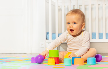 Baby toddler boy building pyramids on the carpet in nursery by the child crib showing surprised engaged face expression