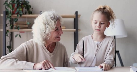 Happy old grandmother helps cute school kid granddaughter dictates writing exercise sitting at table together. Senior grandma teaching grandchild learning handwriting. Two generations studying at home - Powered by Adobe