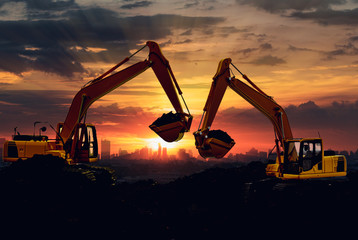 Two Excavators are digging the soil in the construction site on the sky sunset with cityscapes...