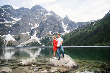 a young couple, a girl in a red dress, a man in a white T-shirt and blue pants stand on a background of mountains near the lake in the park