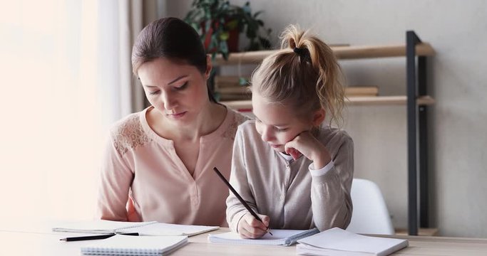 Cute small 6-7 years kid daughter learning writing with young mom tutor. Adult parent mother teaching school child girl helps with homework studying sitting at home table. Children education concept