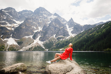 young girl in a red dress stands on a stone against the background of a lake and mountains in the Tatra National Park in Poland