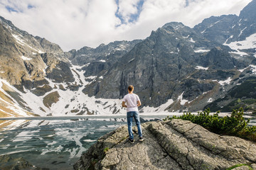 a man in a white T-shirt and blue pants stands on a stone and looks at a lake and mountains in the Tatra National Park in Poland
