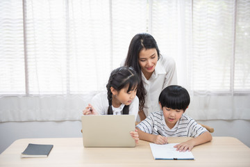 Asian young woman help her son do homework with daughter using laptop sit beside on table in living room at home.