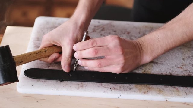 A leather craftsman is making holes in a hand-made belt. The tanner is using a mallet and a hole punch.