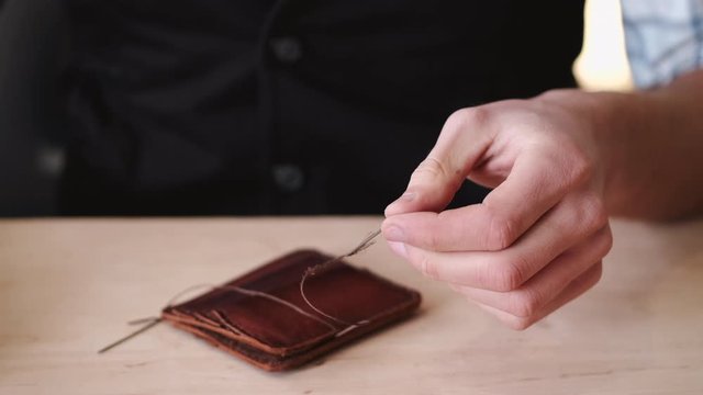 A tanner is stitching a hand-made leather wallet. The currier is using a needle with thread.