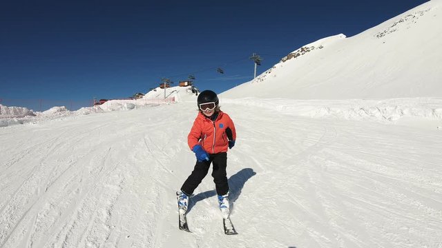 Smiling child skiing and waving hand on snow mountain and blue sky background. Happy boy enjoying winter vacation on ski slopes in sunny day. Having fun with outdoors activity, sport