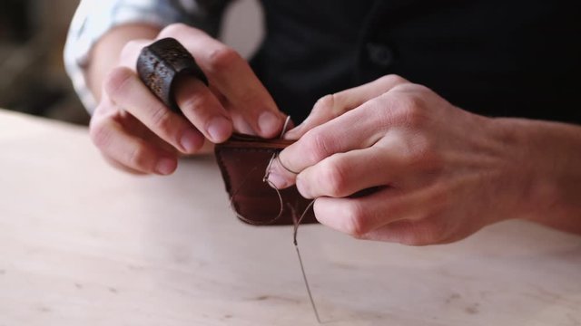 A tanner is stitching a hand-made leather wallet. The currier is using a needle with thread.