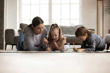 Fototapeta premium Young nanny babysitter lying on floor carpet with small children siblings, spending daytime together at home. Loving caring mother helping daughter and son drawing pictures, developing creativity.