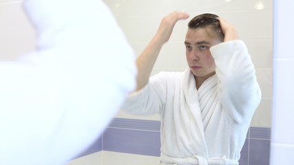 A young man applies mousse for laying on the hair. A man in a white bathrobe in front of a bathroom mirror. View through the mirror