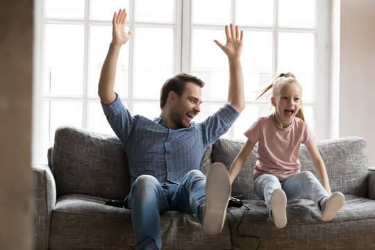 Excited young father raising hands, celebrating video game win with screaming overjoyed little child daughter. Playful small girl enjoying playing xbox with happy laughing dad, weekend hobby time.