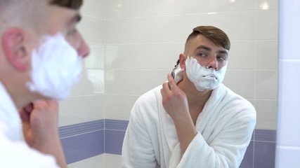 A young man shaves his face hair in front of a mirror. A man in a white coat with foam on his face shaves his hair. View through the mirror.