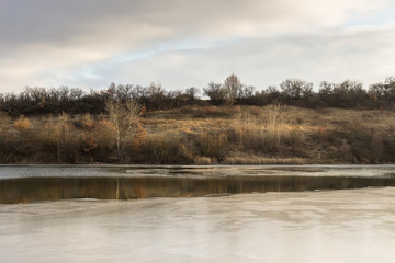 View across beautiful, semi frozen lake with snowflakes on new ice and reed and white birch trees in the forest from the other side