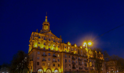 Obraz premium A tall building captured at night. The whole building is lighted with warm yellow light, making it look golden. It contrast with dark navy blue sky. A tall tree growing in front of it.