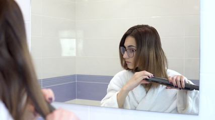 A young woman in glasses straightens her hair with a curling iron in front of a mirror. A girl in a white coat makes a hairstyle in the bathroom. View through the mirror