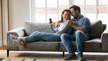 Obraz premium Loving husband cuddling smiling wife, resting together on comfy sofa, using funny mobile applications. Full length overjoyed family couple using smartphone. shopping or watching comedy videos.