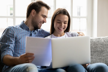 Smiling young man holding papers, embracing happy wife, using computer accounting applications at home. Joyful family couple filling banking information for loan mortgage payment in living room.