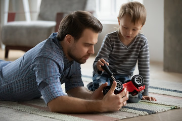 Pleasant young father lying on floor carpet, imagining repairing car with focused little kid son. Small child boy holding screwdriver, fixing broken toy with controlling dad, skills transmission.