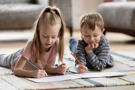 Playful adorable blonde girl lying on carpet floor with younger brother, drawing pictures together in paper album. Interested two little children siblings involved in creative activity hobby at home.
