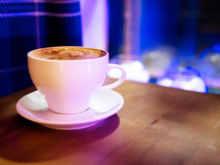 Hot Cappuccino in a cup with froth on wooden table. Coffee shop blue blurred background with bokeh. Mug with milk coffee next to window. Morning and evening beverage concept. Cafe and bar meeting.