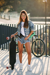 Naklejka na ściany i meble Attractive active woman in sunglasses sits on skateboard at the park area on the cityscape background. Healthy sporty lifestyle concept.