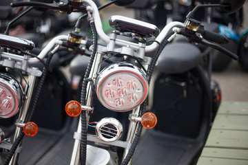 Obraz premium Moto electric vehicles are in a row. Electric bike rental. Headlights at two wheeled vehicles. Motorcycle sale. Mass parking of vehicles. Modern urban way of driving.