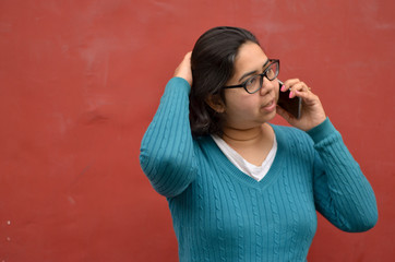 Lifestyle portrait shot of a smart young Indian woman speaking on her mobile phone and running her hands through her hair while standing against a red wall background in New Delhi, India