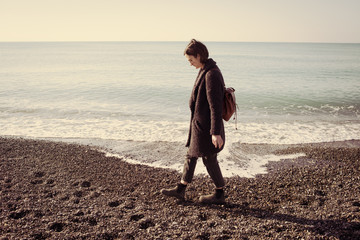 Woman hipster model wearing sunglasses and winter coat walking on a pebble beach with empty calm sea behind. Matte vintage effect.