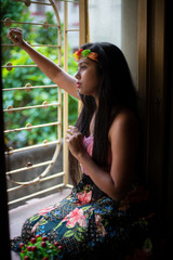 Portrait of an Asian/Japanese/Korean brunette young girl in pink top and skirt and floral headband in front of a window inside of a room. Fashion and boudoir photography. 