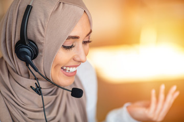 Portrait of happy young islamic woman in headscarf holding microphone and working on computer. Arab businesswoman working in customer service center talking using headset.