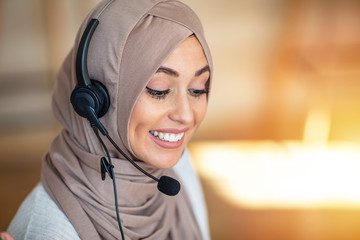 Woman wearing headscarf and headset talking while working in a call centre office, smiling, close-up. Close up beautiful muslim woman call center agent wear headset device and smiling