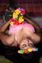 Obraz na płótnie Canvas Top view of an Asian/Japanese/Korean brunette young girl in pink inner wear and floral headband with vibrant flowers lying on bed inside of a room. Fashion and boudoir photography.