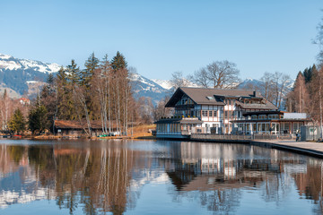 Fototapeta premium Cozy wooden house/chalet on the lake coast. Vacation in Austrian Alps. Snowy mountains in background. Tourism concept. Puddle reflection.