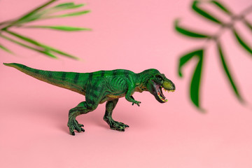 green dinosaur toy with open mouth between blurred palm leaves on a pastel pink  background
