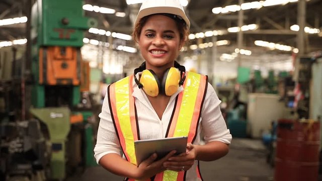 Portrait of professional heavy industry engineer. Technician engineer woman dark skin wearing safety uniform and safety helmet holding digital tablet and smile. Large industrial factory background.