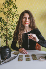 Tyumen, Russia, 12 February 2020 - beautiful young woman in a black velvet dress holds a crystal on a silver chain above the runes during a spiritual seance - obrazy, fototapety, plakaty